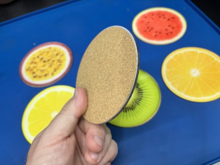 Hand holding a coaster to show its cork backing, with fruit coasters behind